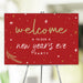 Red New Year’s Eve party welcome sign with gold and white lettering, festive stars, and pine branch accents, ideal decor for holiday celebrations.