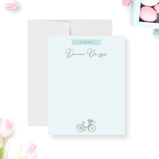 Personalized note card with a pale blue portrait front with a bicycle and flower basket beneath personalized script wording. Shown with a white envelope.