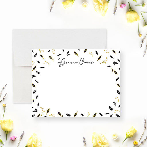 Personalized note card with a white front framed by scattered black and printed gold leaves with a personalized name. Shown with a white envelope.