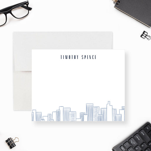 Personalized note card with a white front with blue city skyline line art along the bottom and personalized name at the top. Shown with a white envelope.