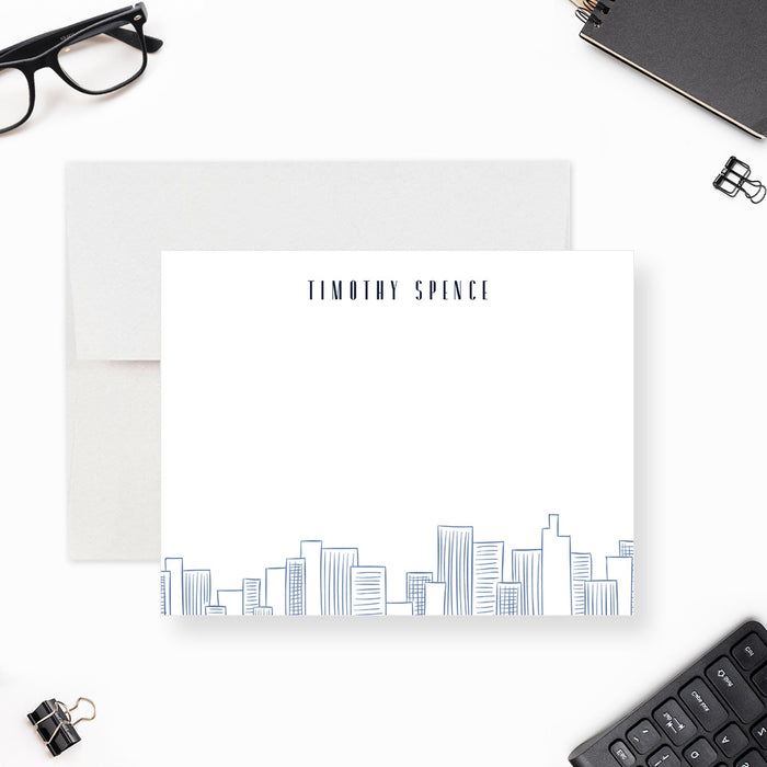 Personalized note card with a white front with blue city skyline line art along the bottom and personalized name at the top. Shown with a white envelope.