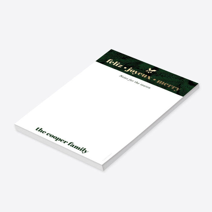personalized holiday notepad with evergreen border, Feliz Joyeux Merry wording, custom family name, and blank writing space-side