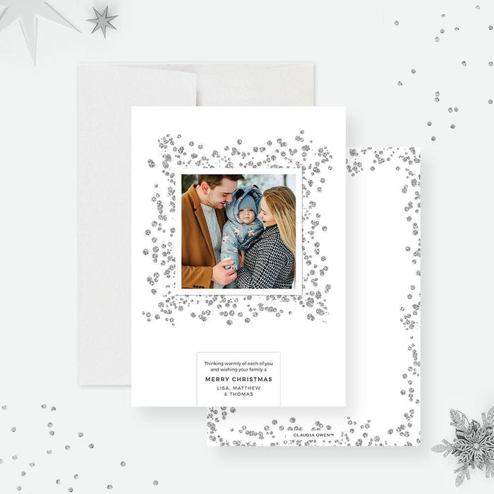 A modern minimalist Christmas photo card featuring a family outdoors in winter, surrounded by silver glitter details and a simple holiday message.