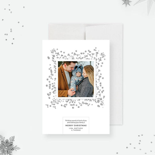 A modern minimalist Christmas photo card featuring a family outdoors in winter, surrounded by silver glitter details and a simple holiday message.