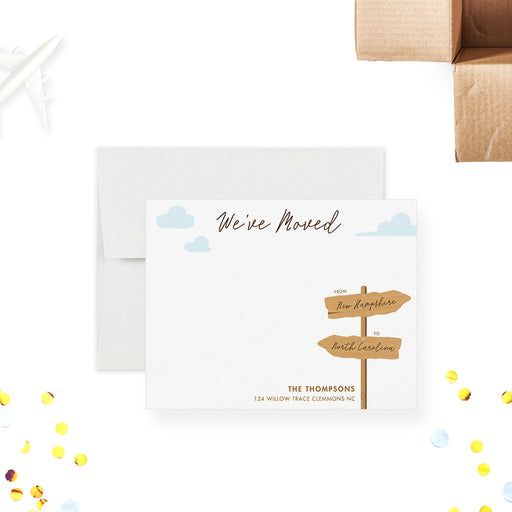 Personalized note card with a wooden moving sign, blue sky, clouds and personalized new address details, shown with a white envelope.