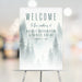 Elegant misty forest wedding welcome sign with personalized names and date, featuring soft pine trees and a romantic woodland theme.