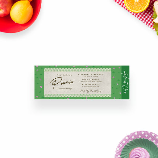 spring picnic ticket invitation with picnic basket illustration, lace border, green grass pattern, tiny pink flowers, and admit one panel