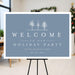 Winter holiday party welcome sign with snowy tree illustration on a blue background displayed on an easel for a Christmas party entrance.